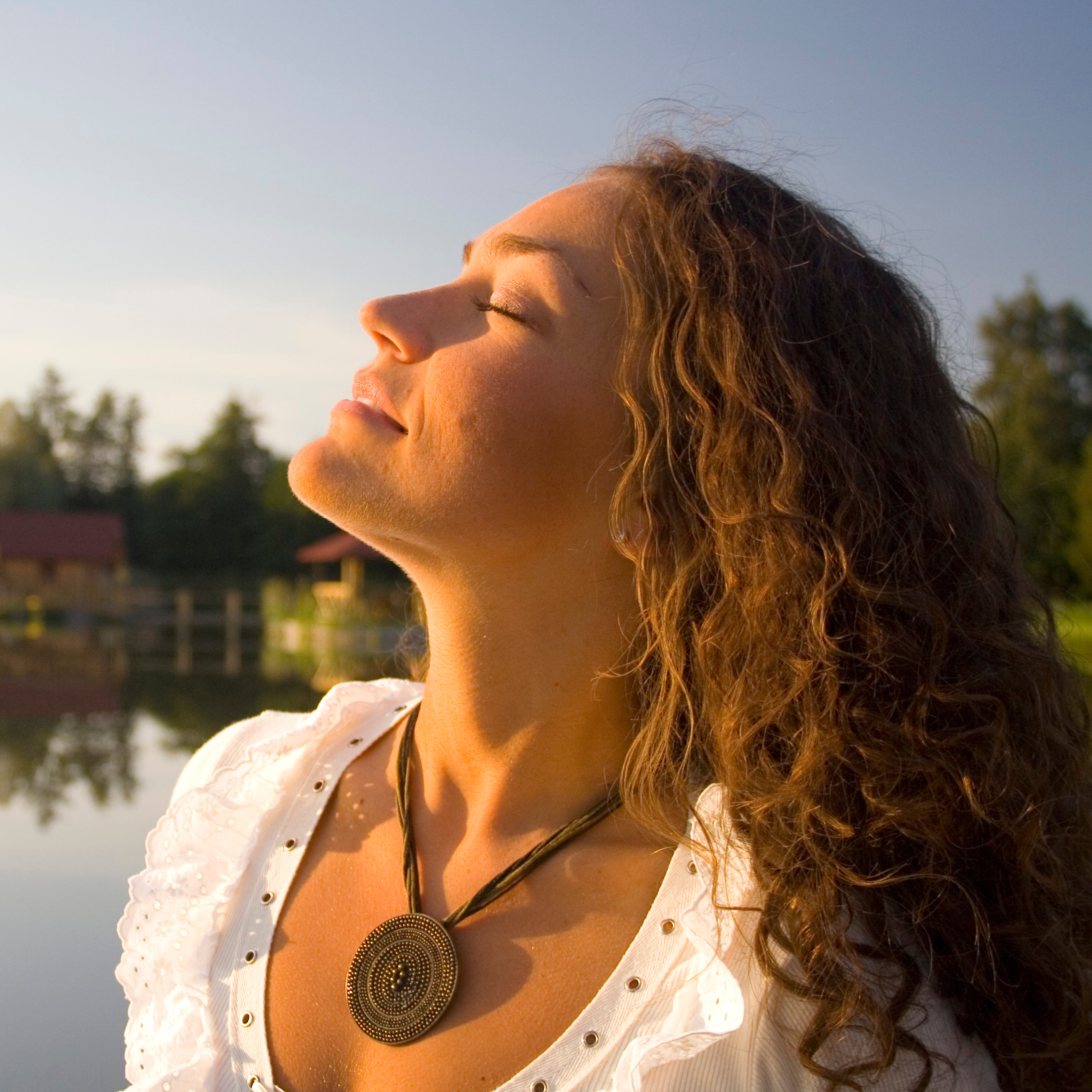En fridfull kvinna med slutna ögon som lutar ansiktet uppåt för att njuta av det varma, naturliga solljuset i en lugn utomhusmiljö vid en sjö.