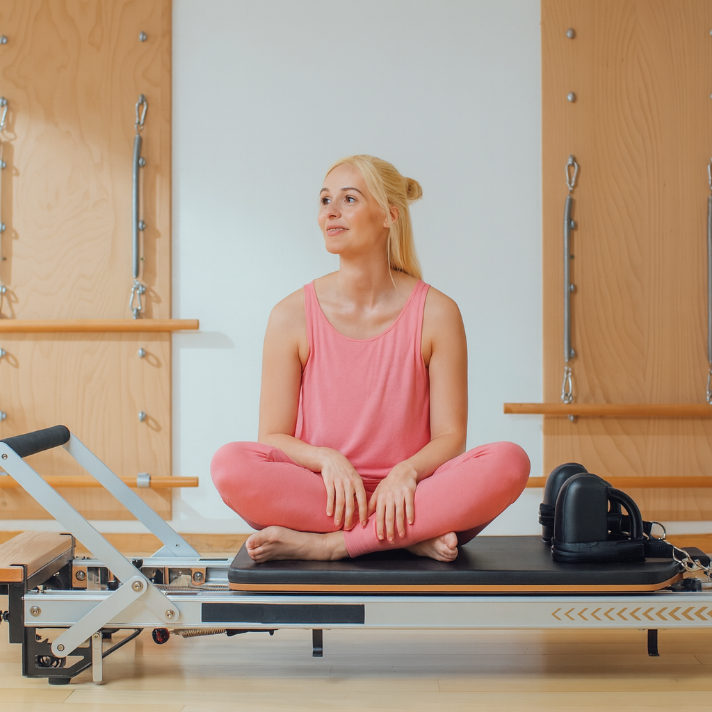 En blond kvinna i rosa träningskläder som sitter fridfullt på en Pilates reformer-maskin efter ett träningspass.